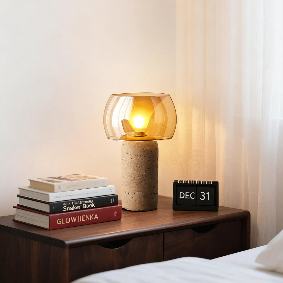 This Clérachic travertine table lamp glowing amber on a walnut bedside chest, next to a stack of books and a flip calendar.