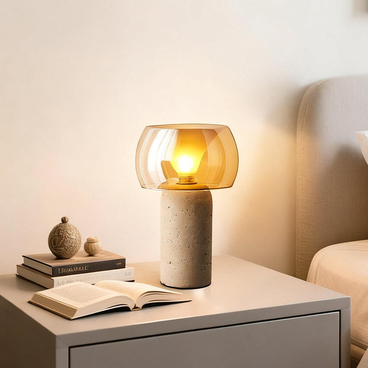 A Clérachic travertine lamp with amber glass shade lit on a pale bedside table, next to an open book and a small ceramic ornament.