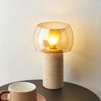 Close, low view of this Clérachic table lamp, its amber shade glowing above a stone column, with a ceramic cup on a dark round table.