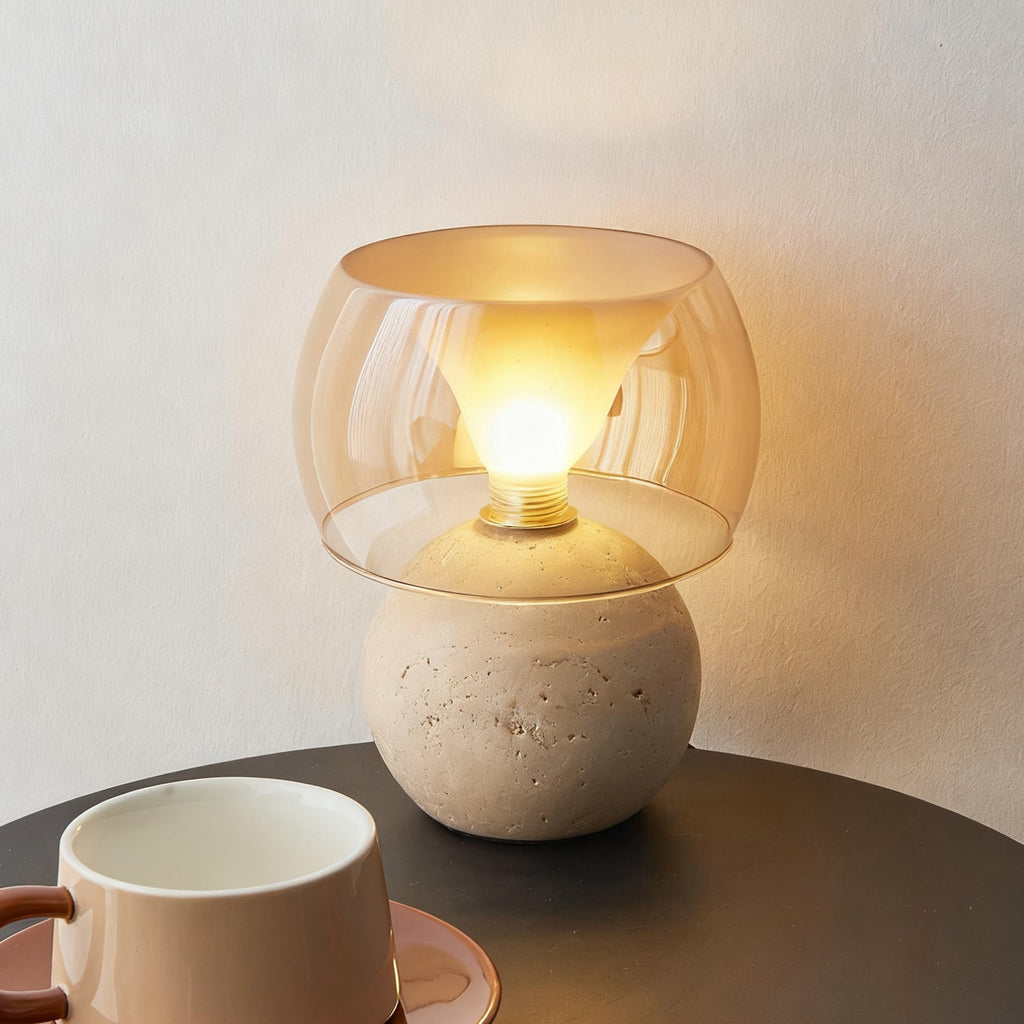 The shorter, rounded-base version of this Clérachic table lamp, lit, with a bulb visible inside the amber glass, beside a cup on a dark table.