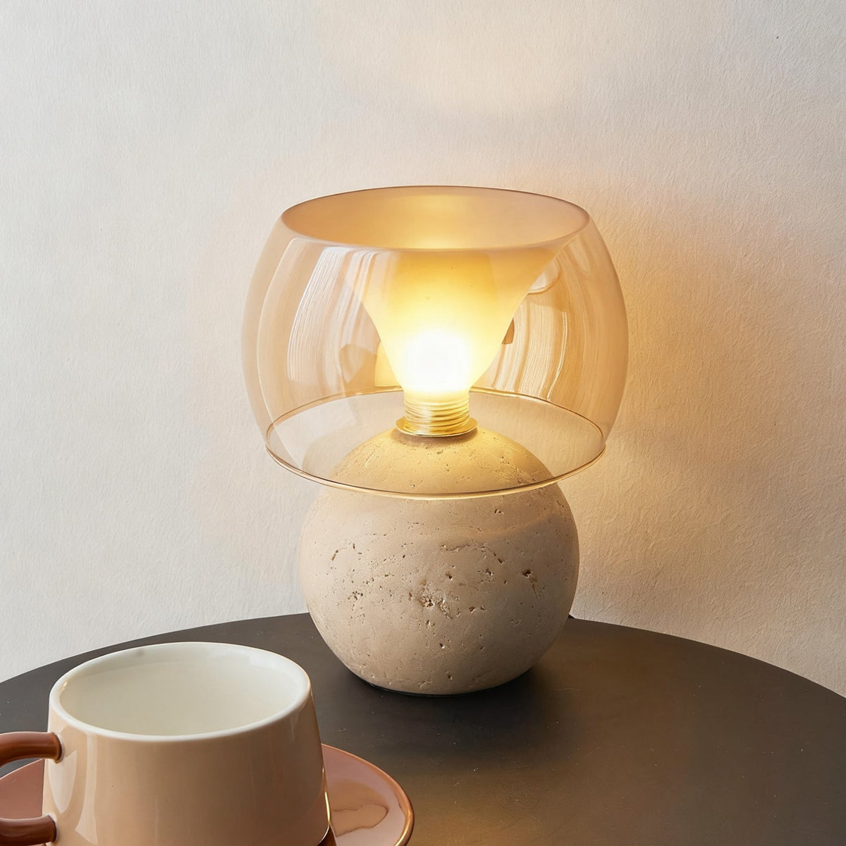 The shorter, rounded-base version of this Clérachic table lamp, lit, with a bulb visible inside the amber glass, beside a cup on a dark table.