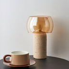 Unlit Clérachic table lamp with an amber glass shade on a travertine column, standing on a dark table beside a cup and saucer.