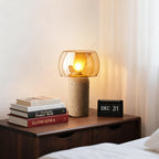 Another view of this Clérachic table lamp lit on a walnut bedside chest, with a flip calendar and books under a sheer window curtain.