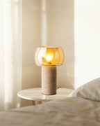 Tall portrait view of a lit Clérachic table lamp on a marble side table, with soft curtain light and bedding at the edge of the frame.