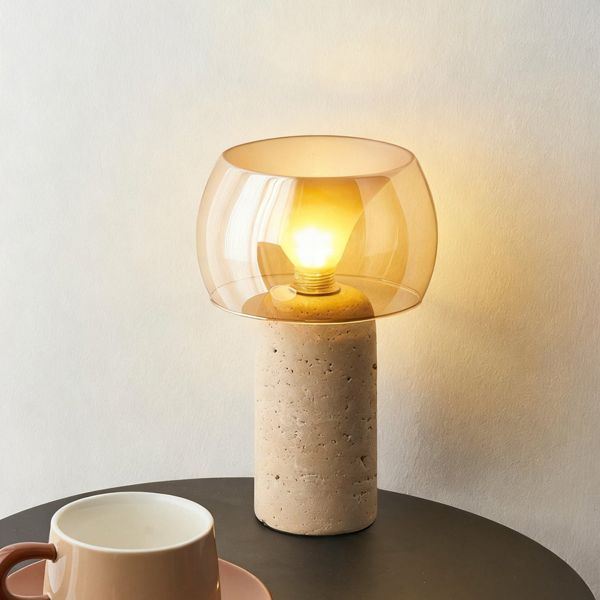 A lit Clérachic table lamp in tight view, its amber shade over a pitted travertine column, with a cup and saucer on a dark table below.