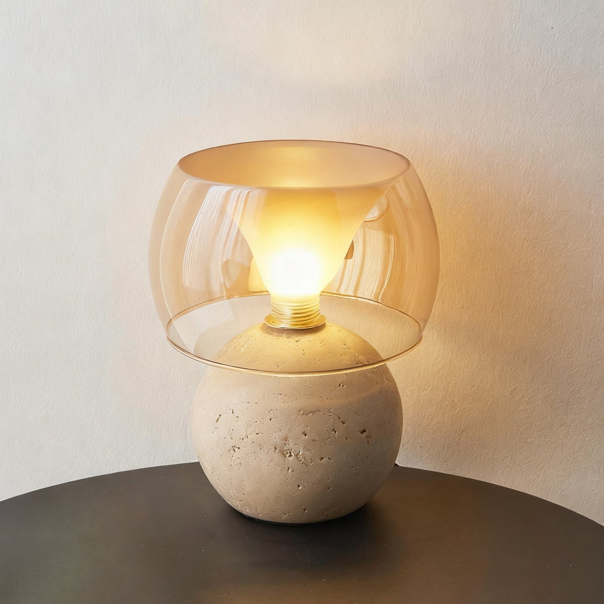 This Clérachic lamp in its rounded-base form, lit, with the bulb glowing inside the amber glass over a pitted travertine sphere.