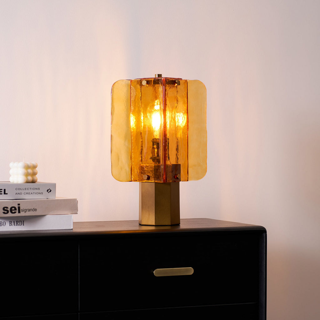 Lit Clérachic table lamp in amber glass on a black chest of drawers, with a small white candle and a stack of books beside it.