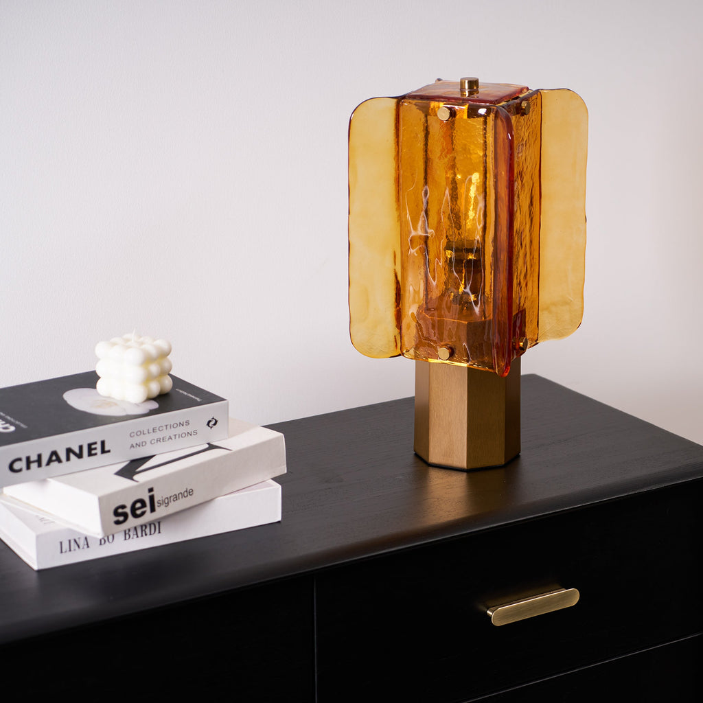 Amber glass panels of this Clérachic table lamp glow on a black sideboard, with stacked books and a bubble candle to the left.