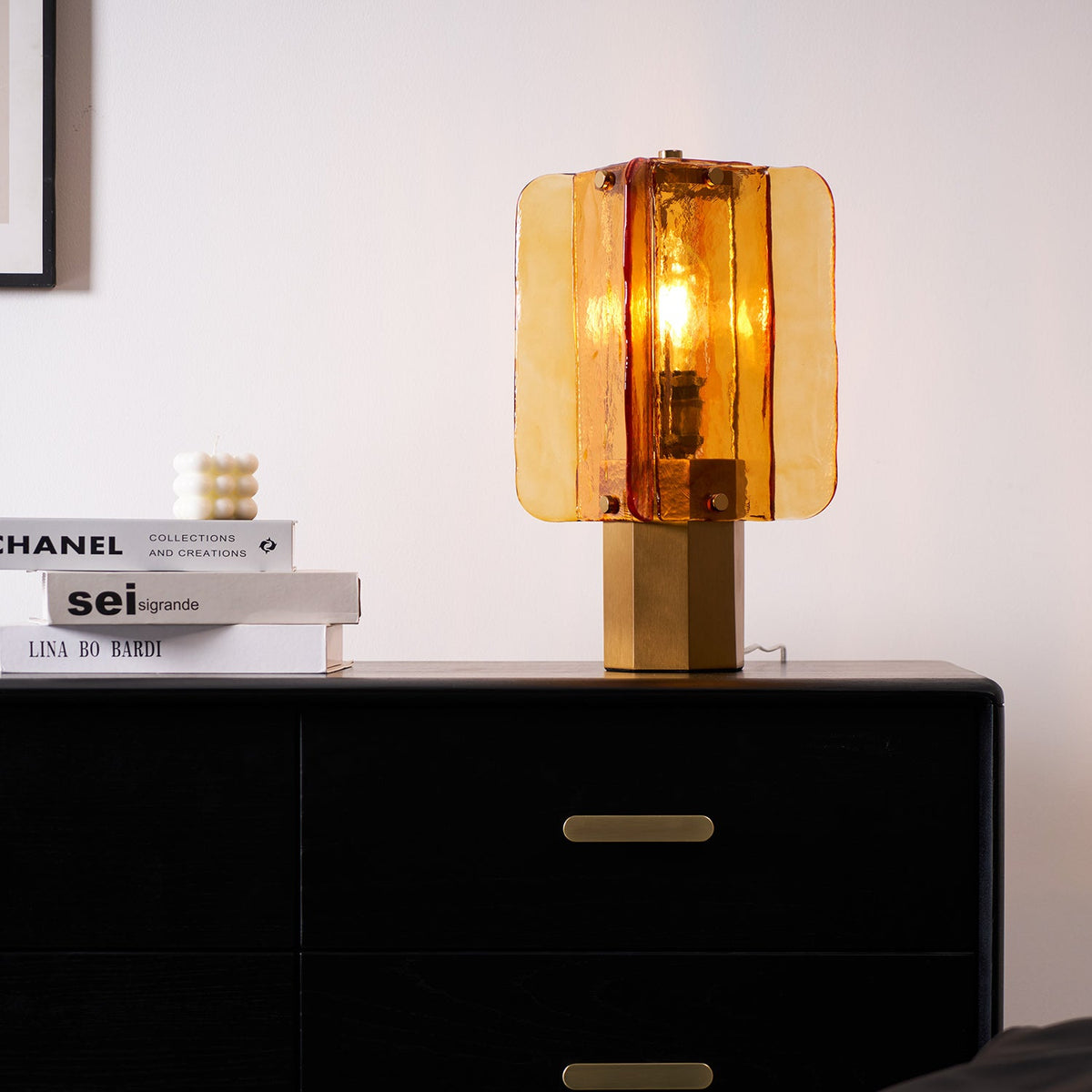 This Clérachic table lamp lights up against a pale wall, standing on a black chest with a brass handle and a stack of books.