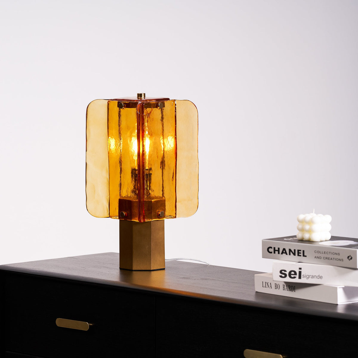 Seen from a low angle, this Clérachic table lamp glows amber on a black cabinet top, with a candle on books at the right.