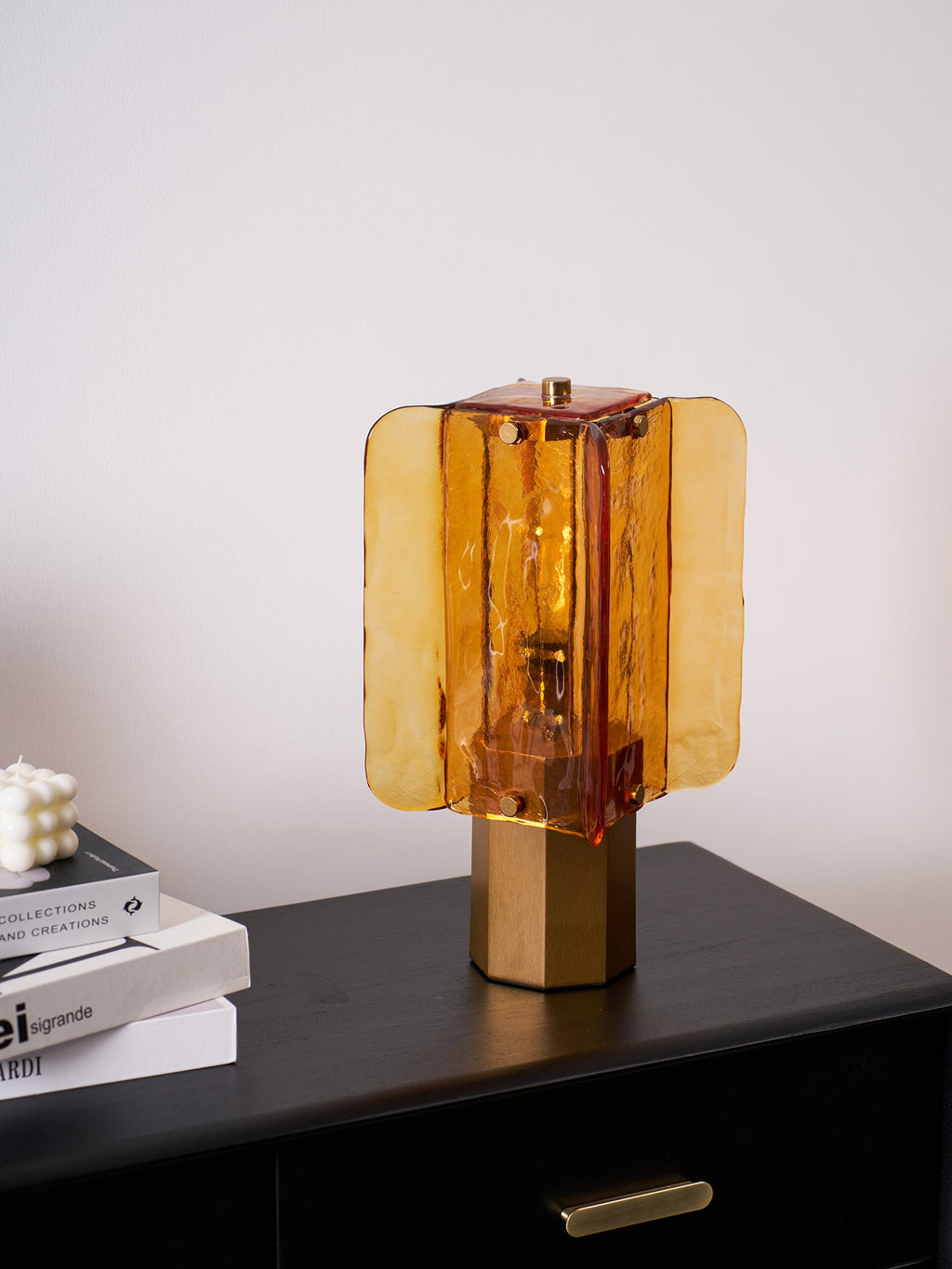 Warm amber light from this Clérachic table lamp on a black chest, with books and a white candle at the left edge of the frame.