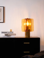 Glowing Clérachic table lamp in amber glass above a black chest of drawers, with a low stack of books and a candle at its left.