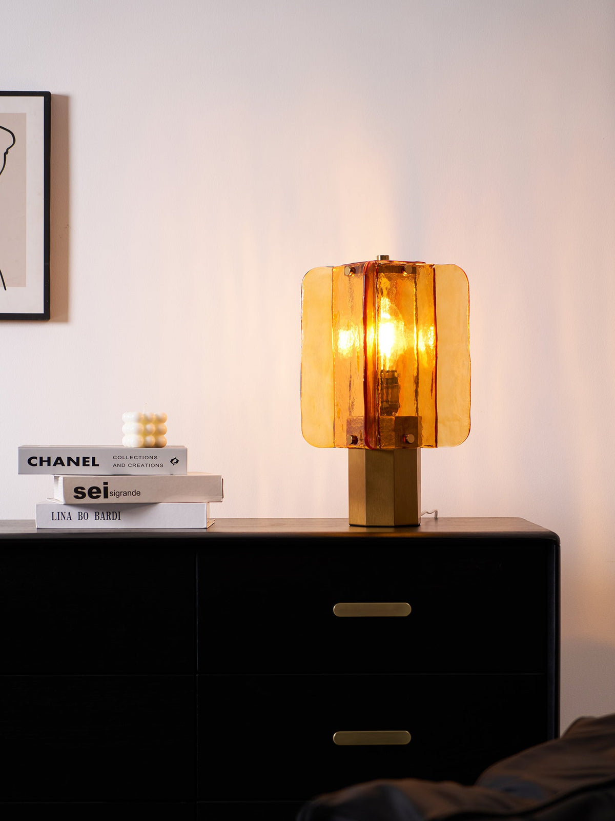 Glowing Clérachic table lamp in amber glass above a black chest of drawers, with a low stack of books and a candle at its left.