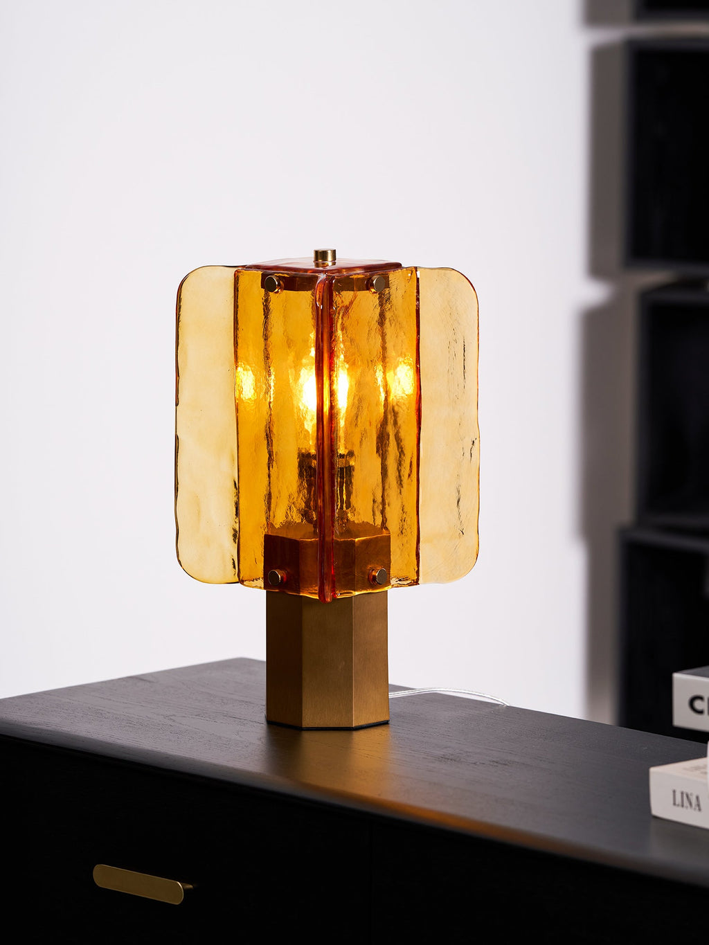 Side view of this Clérachic table lamp in amber glass on a black sideboard, with a hexagonal metal base under the glass panels.