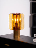 Side view of this Clérachic table lamp in amber glass on a black sideboard, with a hexagonal metal base under the glass panels.