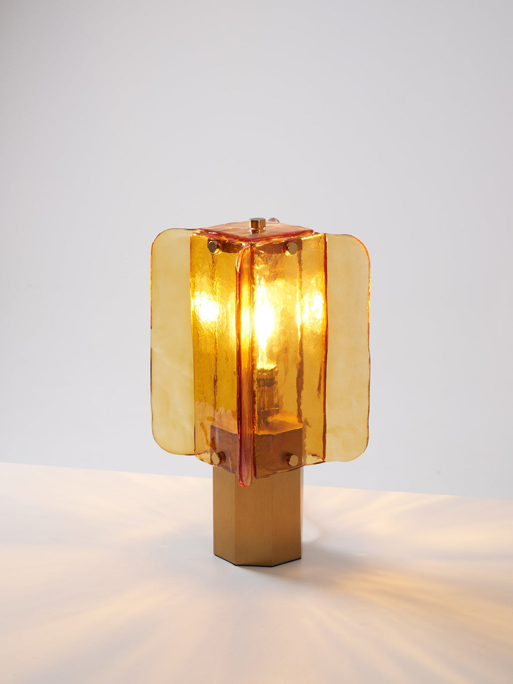 This Clérachic table lamp lit on a white surface, its glow reflecting in soft streaks across the table around the hexagonal base.