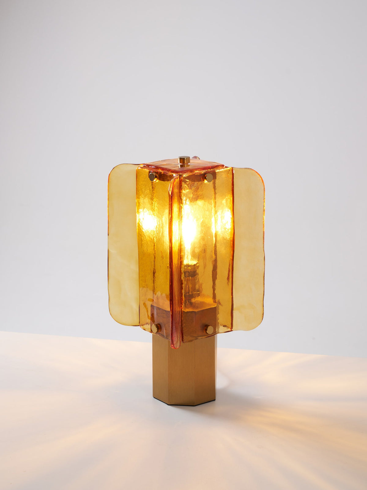 This Clérachic table lamp lit on a white surface, its glow reflecting in soft streaks across the table around the hexagonal base.