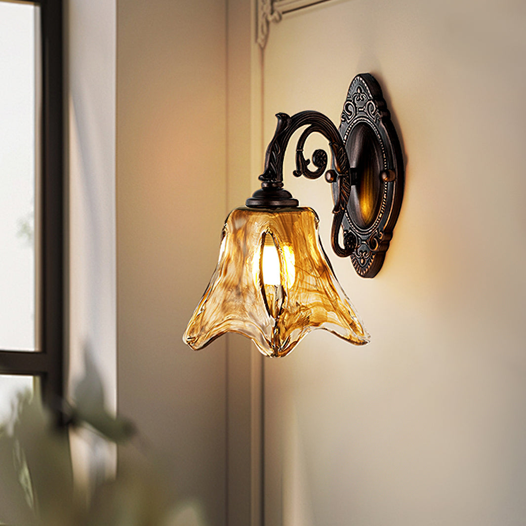Close view of a lit Clérachic wall lamp on a cream wall, its swirled amber shade and ornate scrolled bronze arm clear.