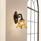 A lit Clérachic wall lamp on a beige wall next to a tall arched window, its amber glass catching the light.