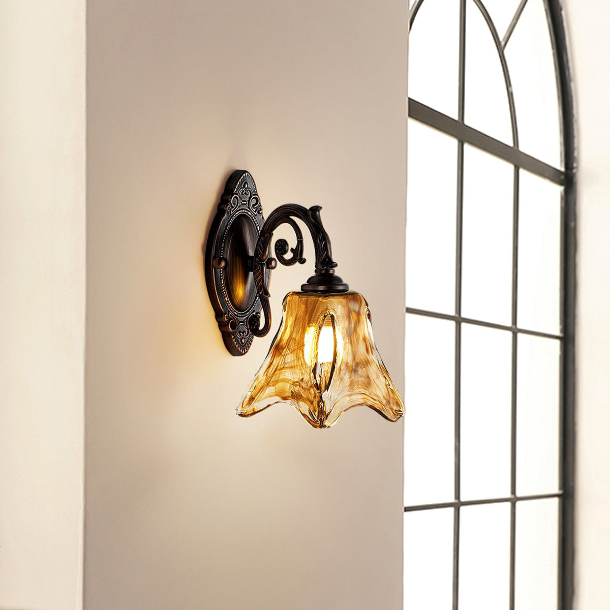A lit Clérachic wall lamp on a beige wall next to a tall arched window, its amber glass catching the light.