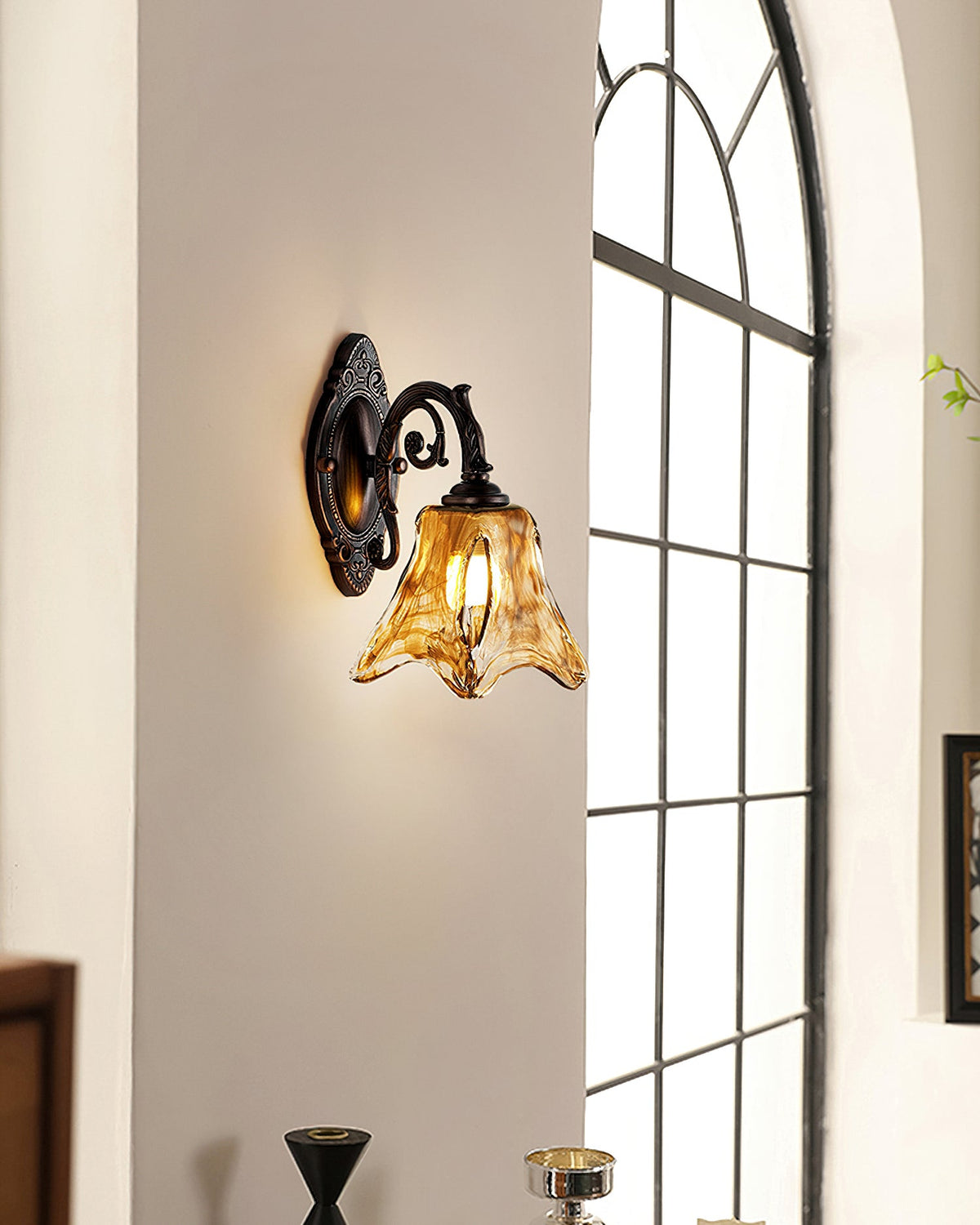 Further back, a Clérachic wall lamp glows on a pale wall beside an arched window with fine black glazing bars.