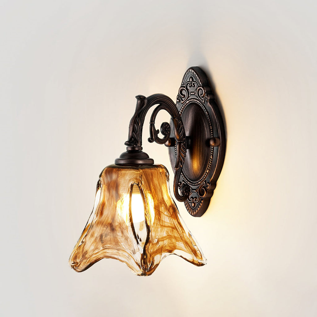 Front view of a Clérachic wall lamp on a pale wall, lit, with the amber glass shade and curling bronze arm.