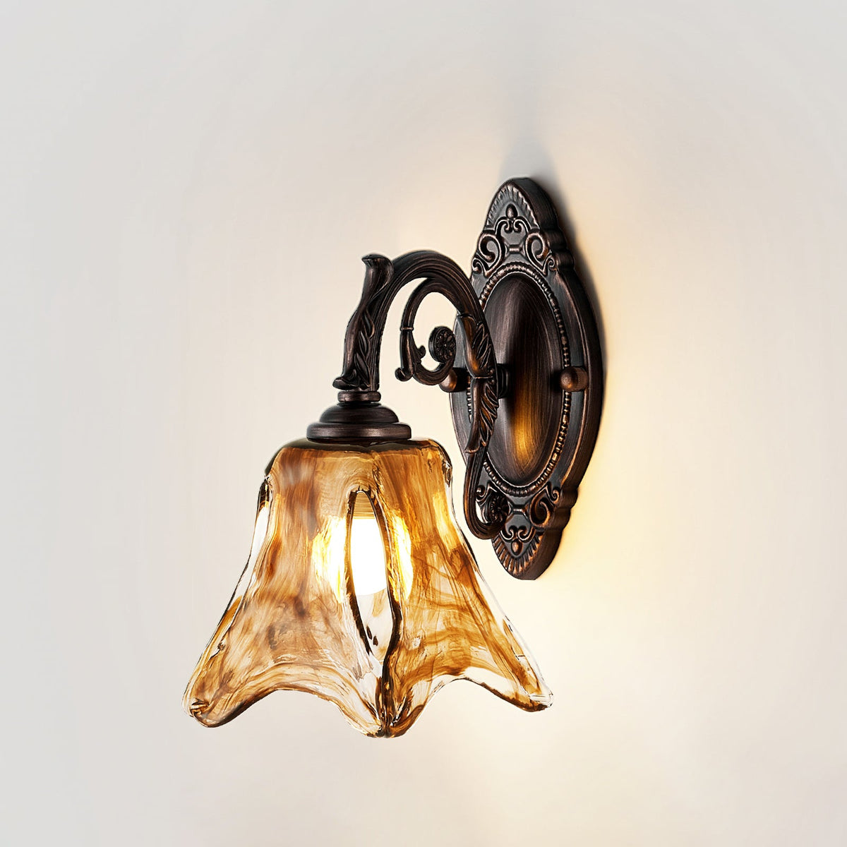 Front view of a Clérachic wall lamp on a pale wall, lit, with the amber glass shade and curling bronze arm.
