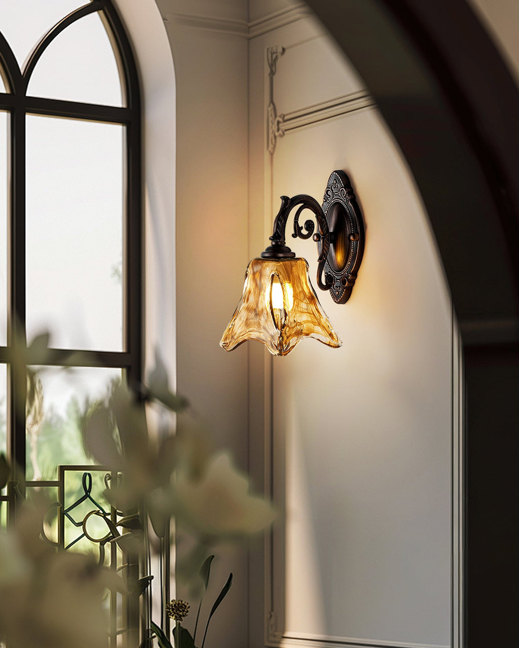 Seen at an angle beside an arched window and a blurred leafy plant, this Clérachic wall lamp glows amber on a cream wall.