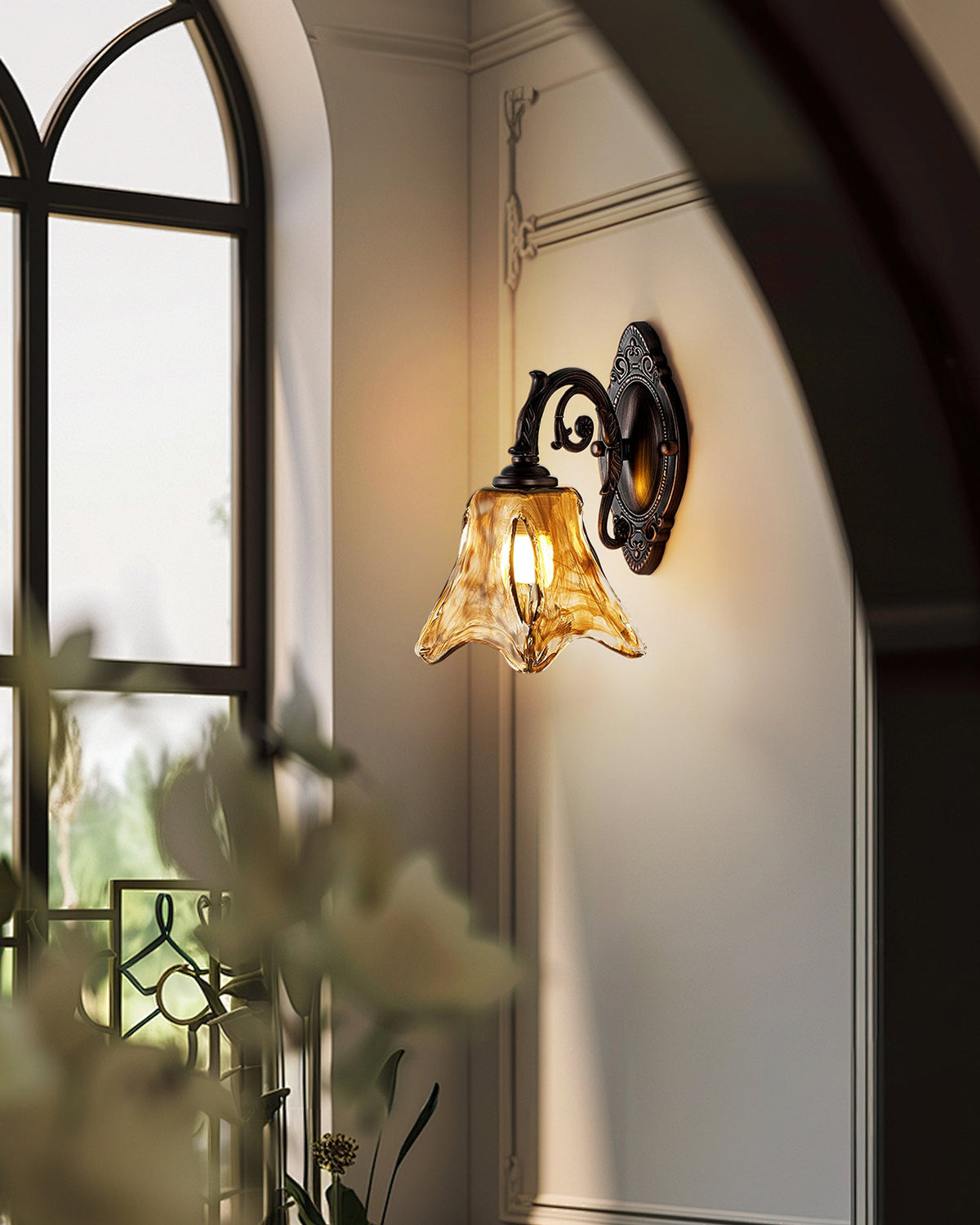 Seen at an angle beside an arched window and a blurred leafy plant, this Clérachic wall lamp glows amber on a cream wall.