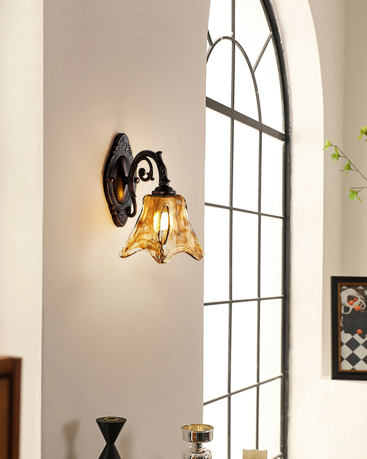 Low in the frame, a lit Clérachic wall lamp sits by an arched window, a black vase and framed picture below.