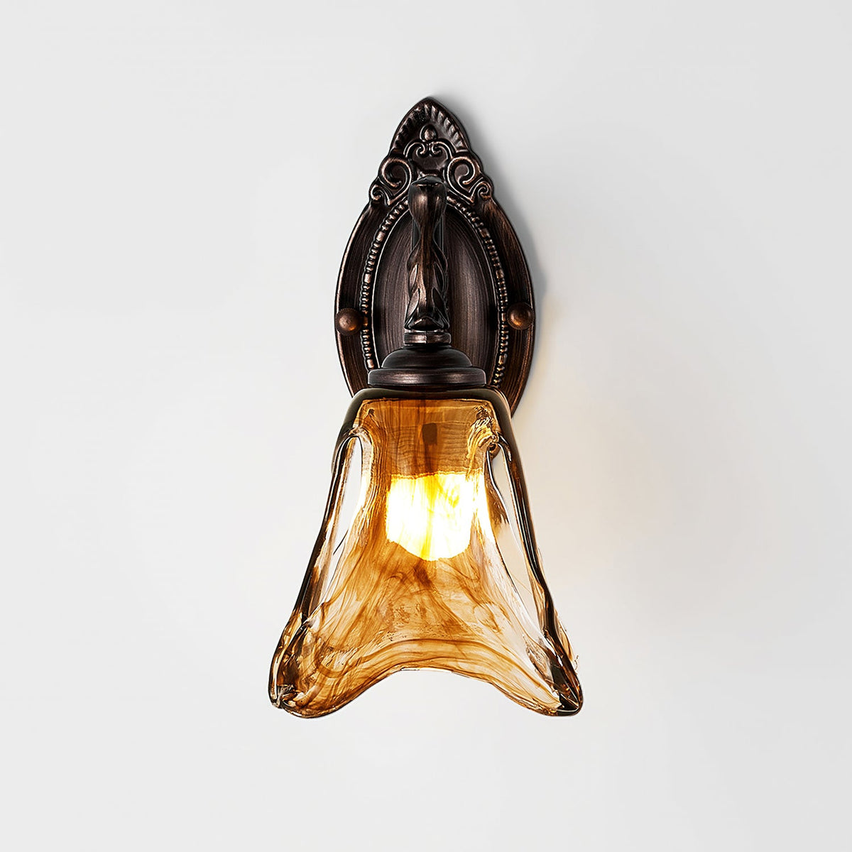 Seen from the front, a Clérachic wall lamp with a narrow ornate backplate and a lit amber glass shade on a pale wall.