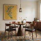 A Clérachic smoky gray glass pendant lights a round dining table beneath a yellow floral painting.