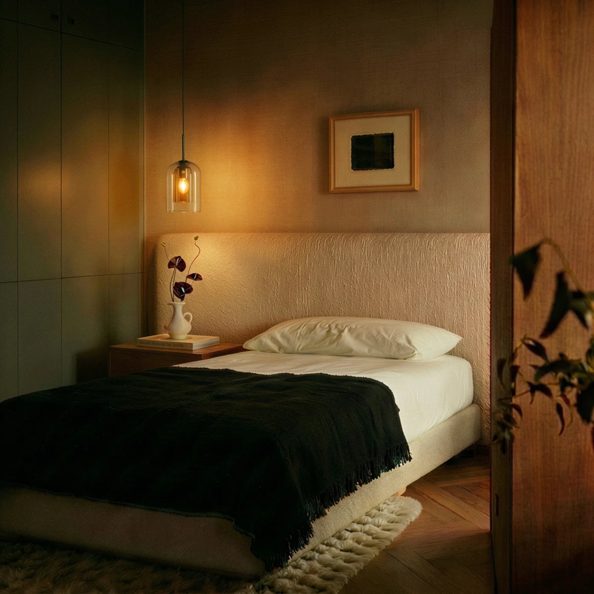 A Clérachic amber glass pendant glows warmly beside a bed, lighting a nightstand with dried flowers.