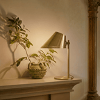 Clérachic table lamp in brass glowing on a mantel beside a carved vase and a leafy potted branch.