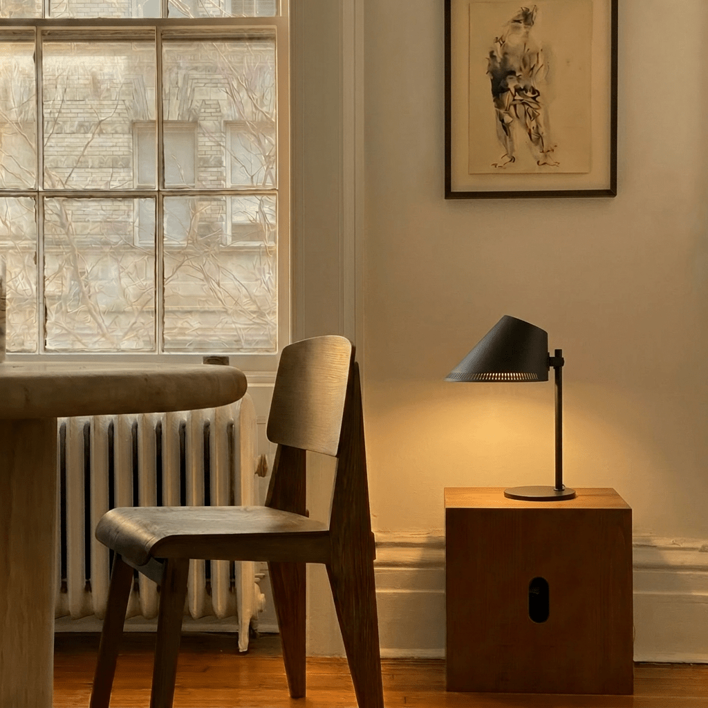 Clérachic table lamp in matte black glowing on a wood block beside a cane chair and framed sketch.