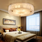 A Clérachic round alabaster ceiling light lit warm above a bed with gold bedding, a tufted headboard and sheer curtains.
