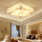 Square alabaster ceiling light from Clérachic lit in a cornice-edged ceiling, with a gold bedhead and framed prints below.