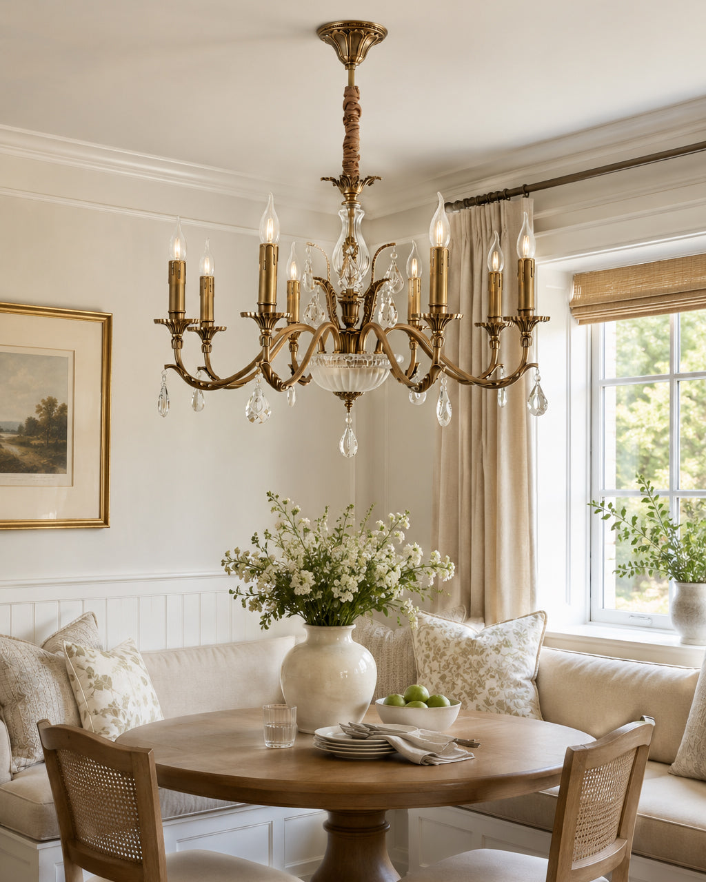 Over a round wooden table and a cushioned corner bench, this Clérachic antique brass chandelier fills a bright, cream-toned room.