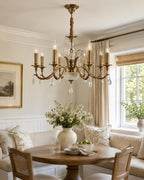 Over a round wooden table and a cushioned corner bench, this Clérachic antique brass chandelier fills a bright, cream-toned room.