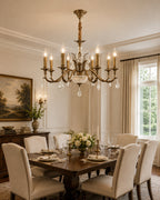 A Clérachic antique brass chandelier lights a dining room with upholstered chairs, a set table, flowers and a framed landscape.