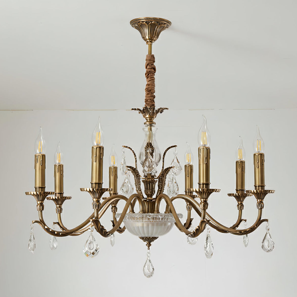 Close view of the brass arms, candle sleeves and crystal drops of this Clérachic chandelier against a pale wall, with its fabric-wrapped rod above.
