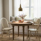 Above a round wooden table by a bright window, a Clérachic brass dome pendant hangs between two cream spindle-back chairs.