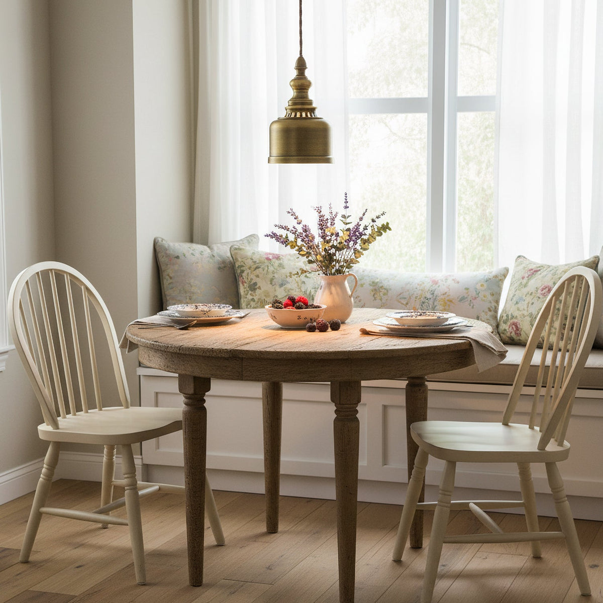 Above a round wooden table by a bright window, a Clérachic brass dome pendant hangs between two cream spindle-back chairs.