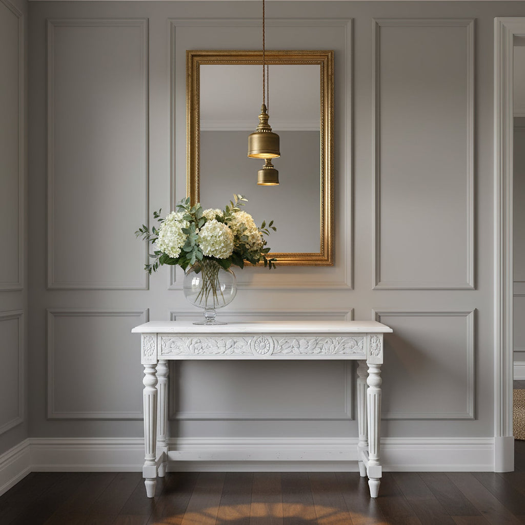 Reflected in a gold-framed mirror, a Clérachic brass dome pendant hangs above a white console table and a vase of white flowers.
