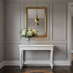 Reflected in a gold-framed mirror, a Clérachic brass dome pendant hangs above a white console table and a vase of white flowers.