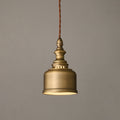 Lit from below, this Clérachic brass dome pendant hangs on a twisted brown cord against a warm grey wall.