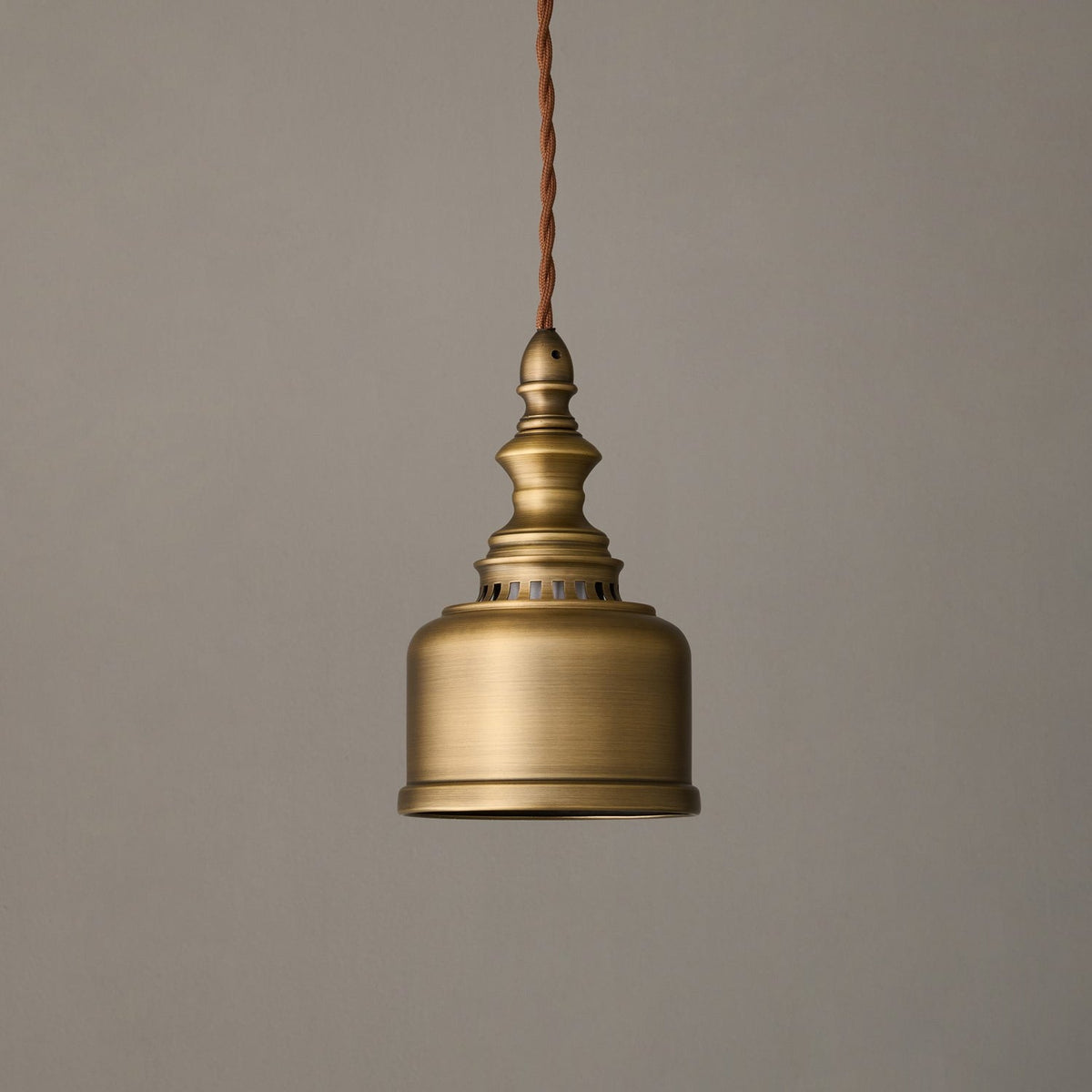Unlit, the antique brass shade and turned finial of this Clérachic pendant hang against a warm grey backdrop.