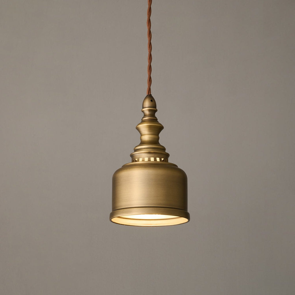 A Clérachic antique brass dome pendant glowing warm on a twisted cord against a soft grey wall, shown at a gentle angle.