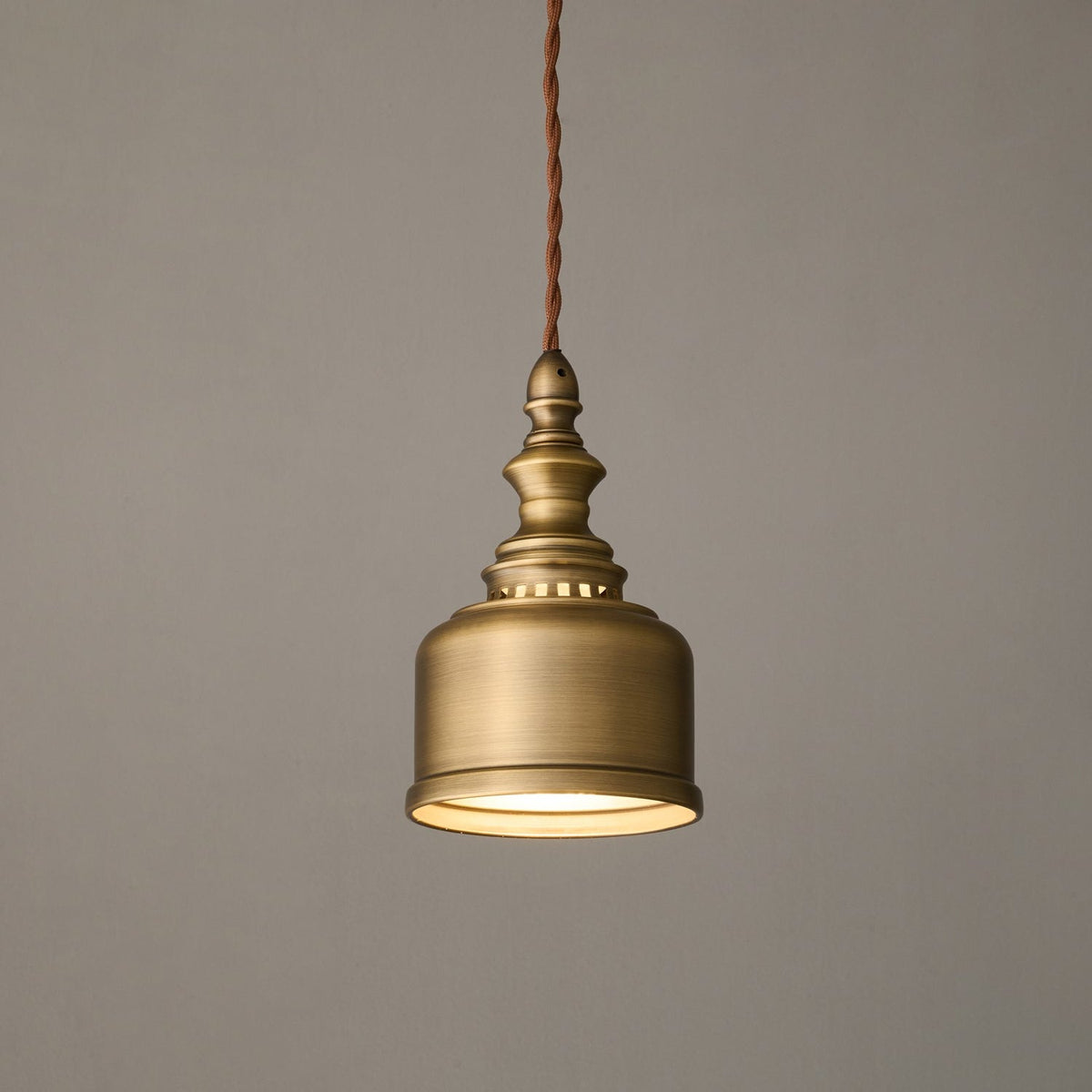 A Clérachic antique brass dome pendant glowing warm on a twisted cord against a soft grey wall, shown at a gentle angle.