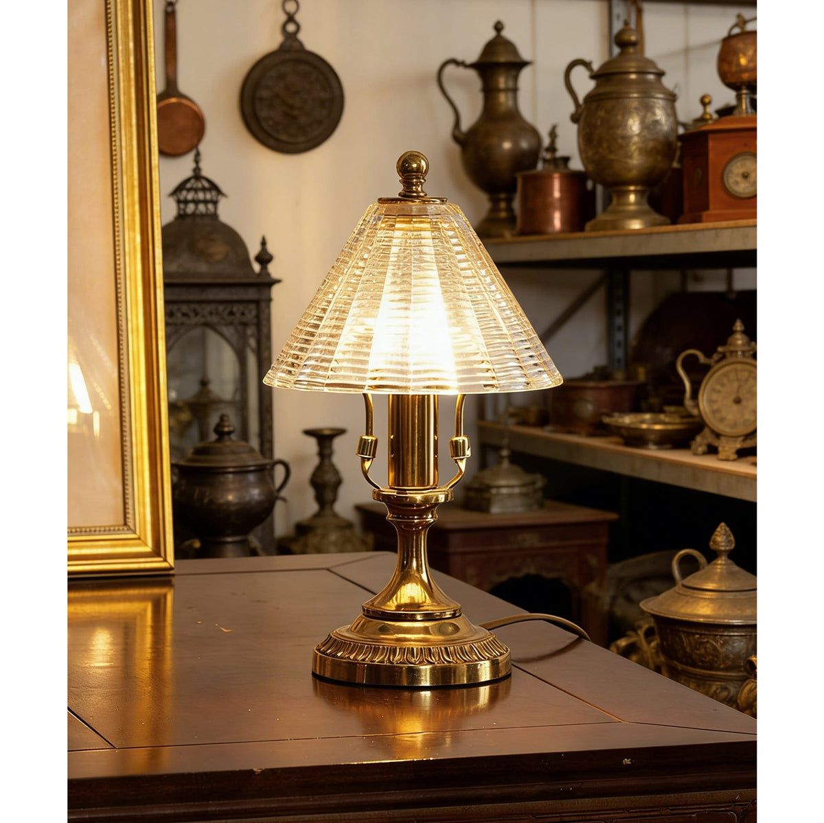 Lit Clérachic table lamp on a dark wooden cabinet, its ribbed glass cone shade glowing warm beside shelves of brassware.